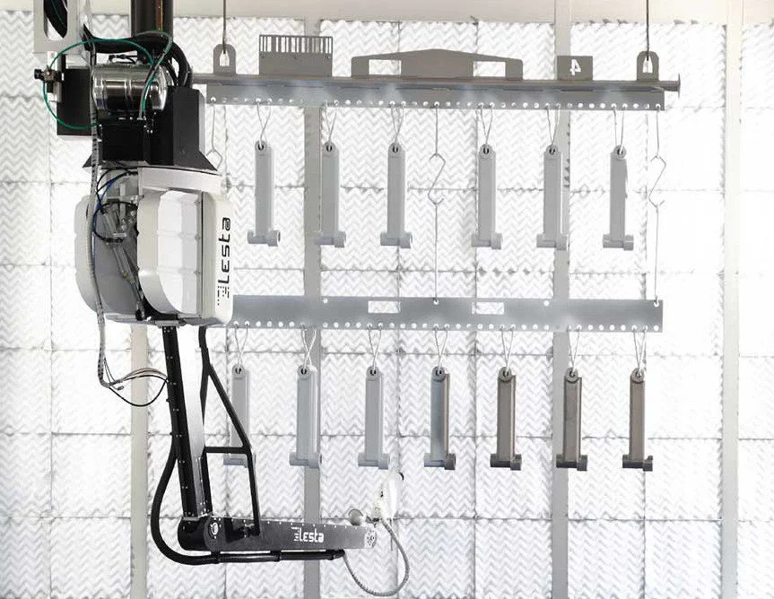A robotic painting arm labeled "Lesta" next to hanging objects in a facility.