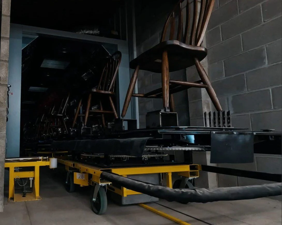 Wooden chairs on a yellow "The PowerCart" system entering a dark chamber replacing outdated conveyor systems with autonomous, explosion-proof carts.